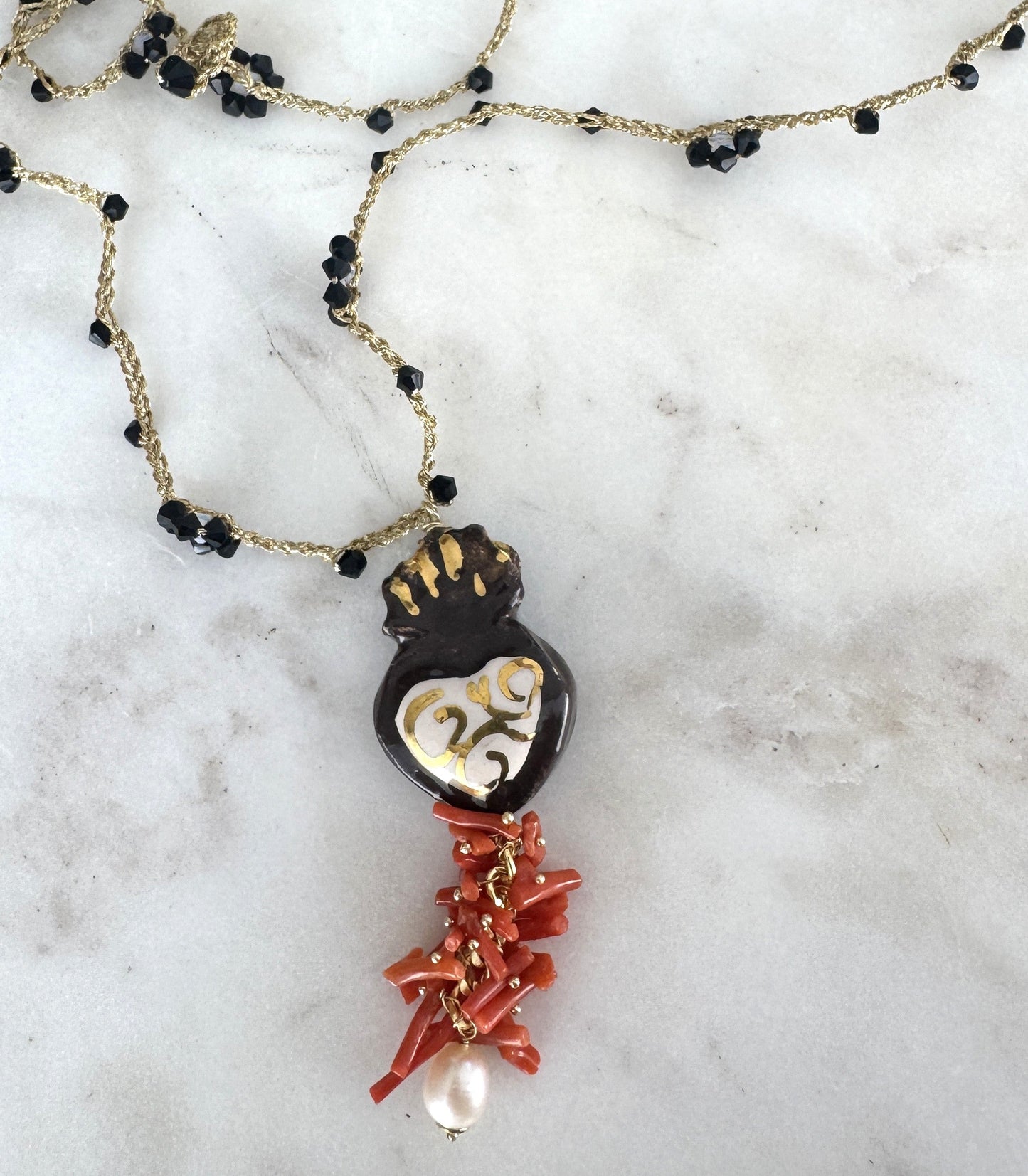 Hecho en Italia, el Collar Cuore Sacro combina cerámica artesanal con detalles dorados, coral rojo natural, perla barroca y cristales negros sobre cadena bañada en oro. Una joya simbólica y poderosa que refleja arte, historia y sofisticación mediterránea.