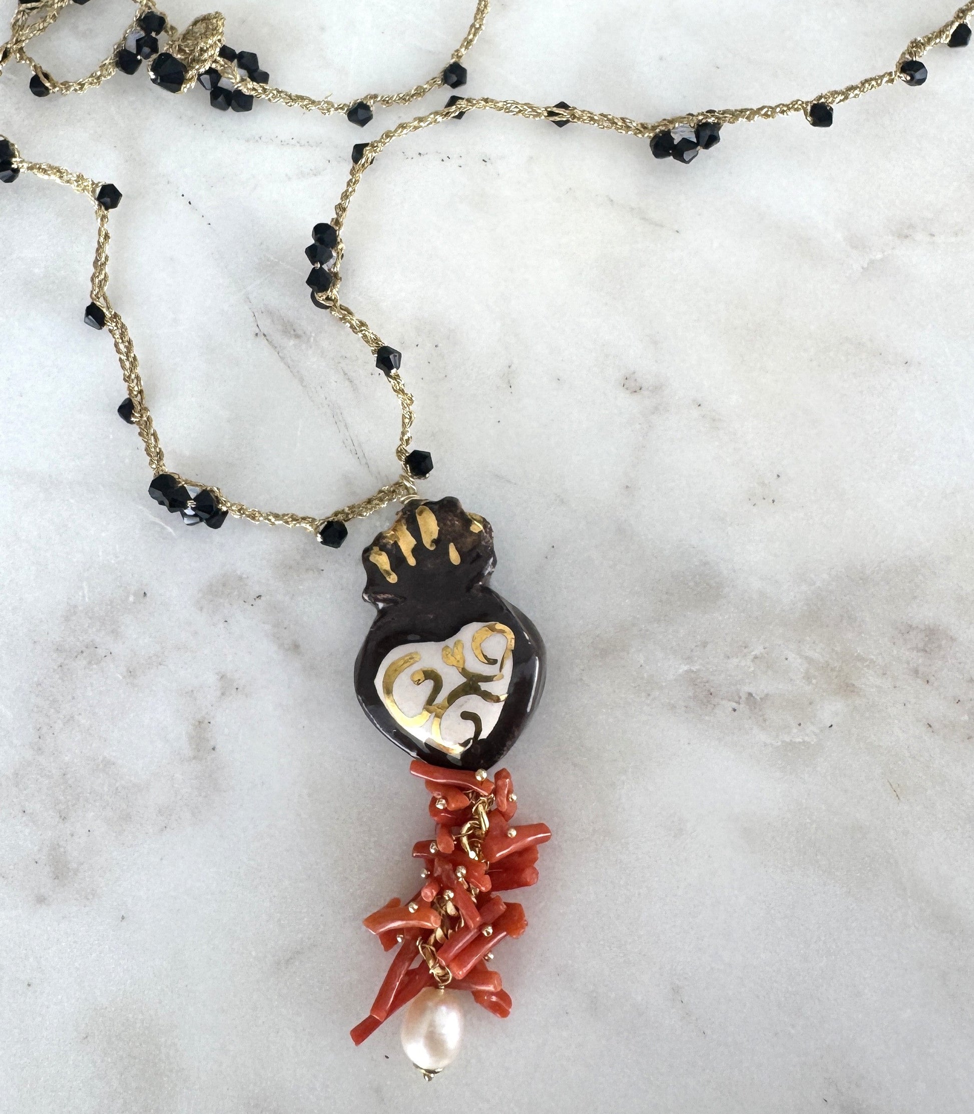 Hecho en Italia, el Collar Cuore Sacro combina cerámica artesanal con detalles dorados, coral rojo natural, perla barroca y cristales negros sobre cadena bañada en oro. Una joya simbólica y poderosa que refleja arte, historia y sofisticación mediterránea.