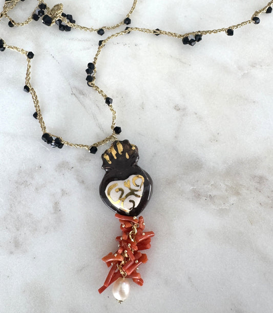 Hecho en Italia, el Collar Cuore Sacro combina cerámica artesanal con detalles dorados, coral rojo natural, perla barroca y cristales negros sobre cadena bañada en oro. Una joya simbólica y poderosa que refleja arte, historia y sofisticación mediterránea.